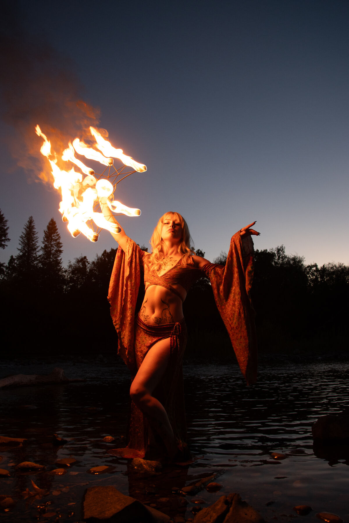 Fire performer standing in a river at dusk with fire fans raised