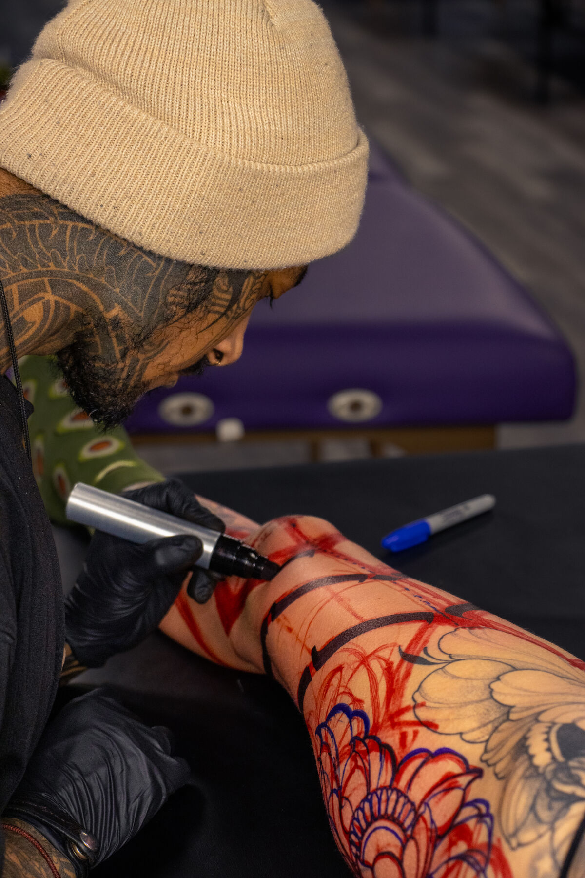 Tattoo artist in a cream beanie drawing a stencil on a client's leg